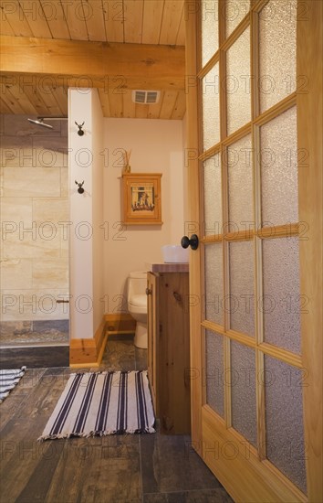 Opened frosted glass French door of main bathroom with tan and dark grey ceramic tile floor and beige ceramic tile shower stall, blue and white striped rug on ground floor inside luxurious contemporary timber and milled log cabin home, Quebec, Canada, North America