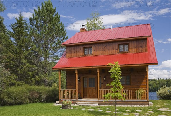 Small two story brown stained log cabin home with veranda and red sheet metal roof and landscaped front yard with deciduous tree and flagstone footpath in summer, Quebec, Canada, North America