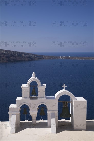 White roughcast cladded arched church bells tower with cross overlooking the Aegean sea, Oia village, Santorini, Greece, Europe