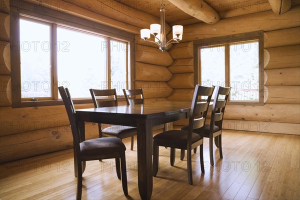 Dark brown stained wooden table and upholstered seat high back chairs in dining room with illuminated chandelier and varnished floor boards inside luxurious contemporary Scandinavian style log cabin home, Quebec, Canada, North America