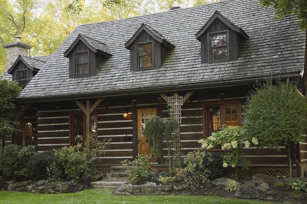 White chinked log cabin home facade with grey weathered cedar shingles roof, three dormer windows and landscaped front yard with mixed perennial shrubs and plants in autumn, Quebec, Canada, North America