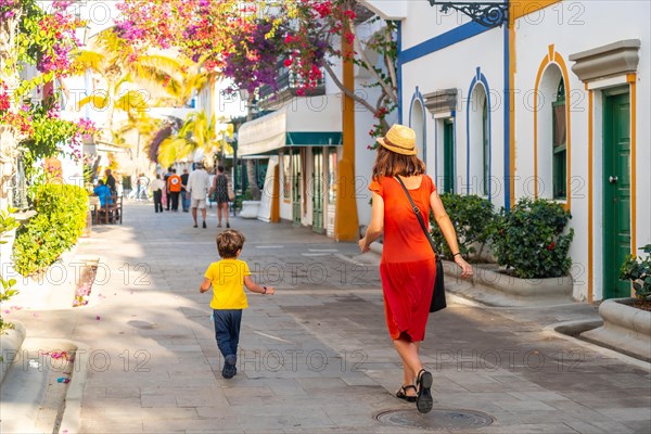 A woman and a child walk down a street with a lot of shops. The woman is wearing a red dress and a straw hat