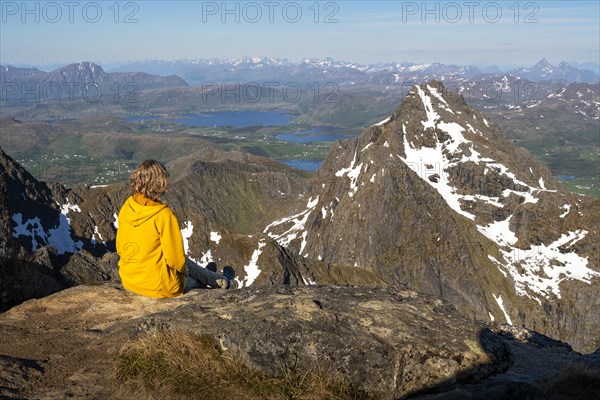 Mountain landscape in the Lofoten Islands. A hiker sits on the summit of Himmeltindan mountain and looks out over the landscape. Good weather, sunshine and blue sky. Vestvagoya, Lofoten, Norway, Europe