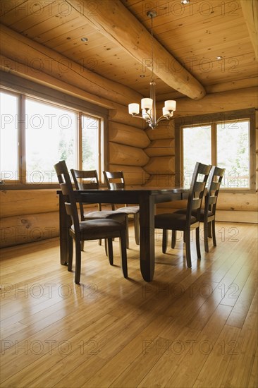 Dark brown stained wooden table and upholstered seat high back chairs in dining room with illuminated chandelier and varnished floor boards inside luxurious contemporary Scandinavian style log cabin home, Quebec, Canada, North America