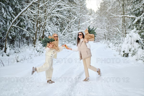 Two joyful women in snowy forest holding fir tree branches on their shoulders and clinking glasses with champagne