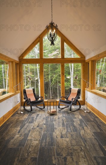 Reading room with tan and dark grey nuanced ceramic tile floor and two black leather rocking chairs plus pendant medieval candle light bulb style black wrought iron chandelier inside luxurious contemporary timber framed and milled log cabin home, Quebec, Canada, North America