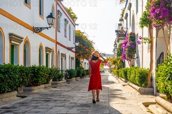 A woman in a red dress walks down a narrow street with a hat on. The street is lined with trees and houses, and there are potted plants along the sidewalk. The scene has a peaceful