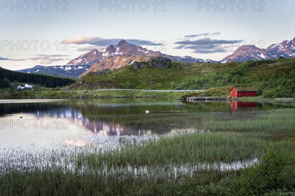Landscape in the Lofoten Islands. Lake Holdalsvatnet with vegetation on the shore. A red wooden house. Mount Blatinden and other mountains in the background. The landscape is reflected in the lake. At night at the time of the midnight sun in good weather, blue sky. Vestvagoya, Lofoten, Norway, Europe