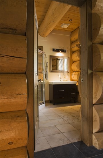 Wooden vanity with white rectangular sink in illuminated guest bathroom with tan ceramic tile floor viewed through doorway in log wall inside luxurious contemporary Scandinavian style log cabin home, Quebec, Canada, North America