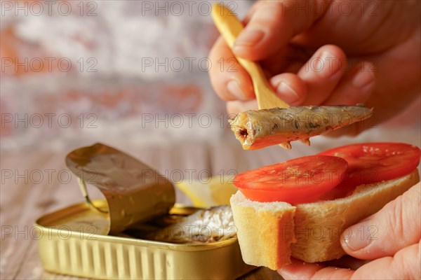 A woman puts a sardine on a slice of bread with tomato, in the background you can see the can of sardines and a lemon on a wooden table