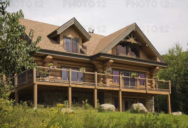 Rear of large two story luxurious Scandinavian style log cabin home with timber design elements and long elevated patio deck, a balcony with black wrought iron railing and brown and tan asphalt shingles roof in summer, Quebec, Canada, North America