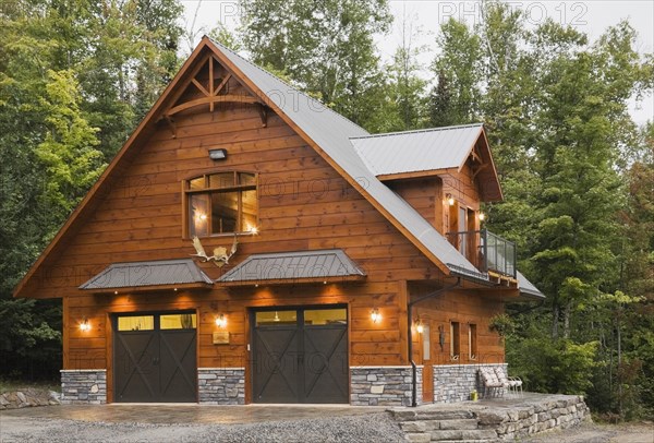 Illuminated milled Eastern white pine knotty wood plank two story flat log profile and timber two door garage building with grey nuanced natural stone cladding and sheet metal roof in early evening light in late summer, Quebec, Canada, North America