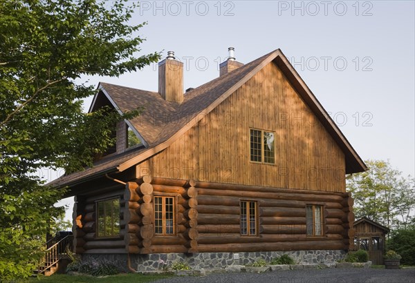 Facade and side view of rustic contemporary Scandinavian hybrid style log cabin home with light brown stained vertical wood plank siding, two tone asphalt shingles roof and chrome top chimneys in summer, Quebec, Canada, North America