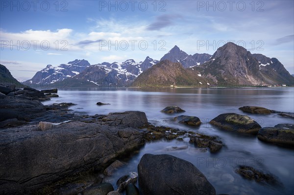 Landscape with sea and mountains on the Lofoten Islands, view over the fjord Flakstadpollen to the small village Flakstad and the mountains Flakstadtinden, Stortinden and other mountains. Rocks in the foreground. At night at the time of the midnight sun in good weather, some clouds in the sky. Long exposure. Early summer. Flakstadoya, Lofoten, Norway, Europe