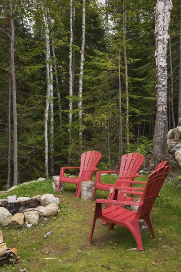 Four bright red plastic Adirondack chairs around rock edged firepit dug into the ground in backyard surrounded by a forest of evergreen and deciduous trees in late summer, Quebec, Canada, North America