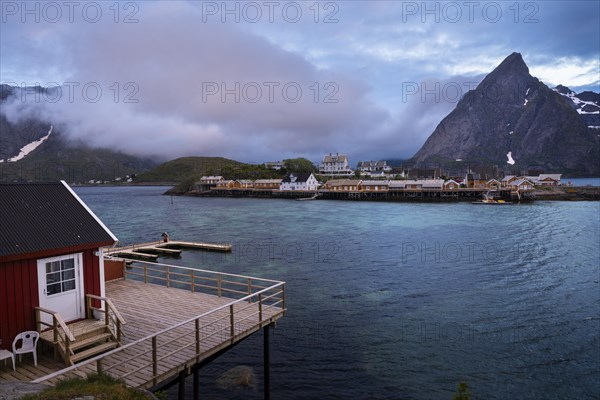 View of Sakrisoy with its typical yellow or ochre-coloured wooden houses by the sea, with Mount Olstinden in the background. In the foreground, a typical red wooden house (rorbuer) with a terrace on wooden stilts. At night at the time of the midnight sun. A few clouds in the sky. Early summer. Sakrisoy, Moskenesoya, Lofoten, Norway, Europe