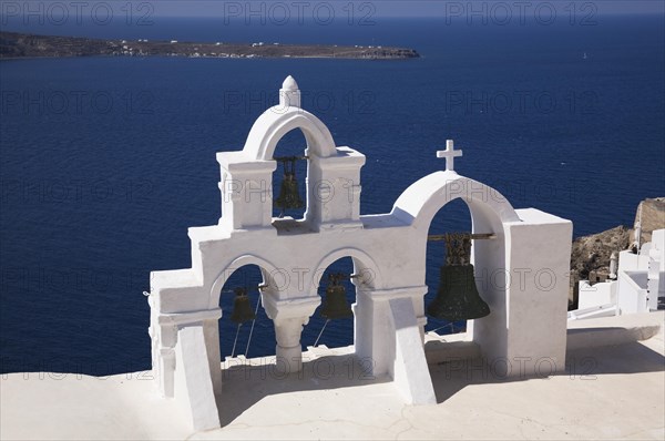 White roughcast cladded arched church bells tower with cross overlooking the Aegean sea, Oia village, Santorini, Greece, Europe
