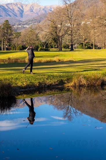 Male Golfer Reflected in a Water Pond and Hitting the Golf Ball on Fairway on Golf Course with Mountain in a Sunny Day in Switzerland. | MR:yes Mats-CH-02-05-2023