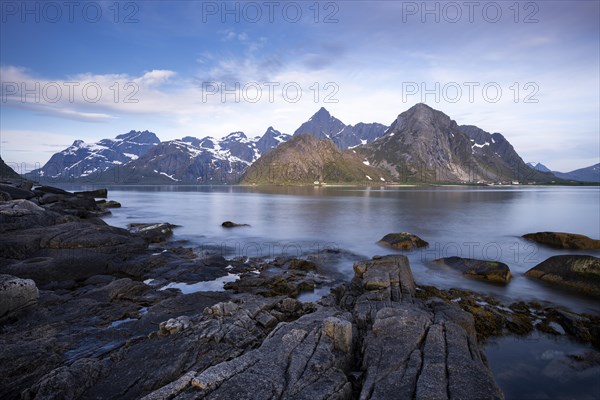 Landscape with sea and mountains on the Lofoten Islands, view across the fjord to the small town of Flakstad and the mountains Flakstadtinden, Stortinden and other mountains. Rocks in the foreground. At night at the time of the midnight sun in good weather, some clouds in the sky. Long exposure. Early summer. Flakstadoya, Lofoten, Norway, Europe