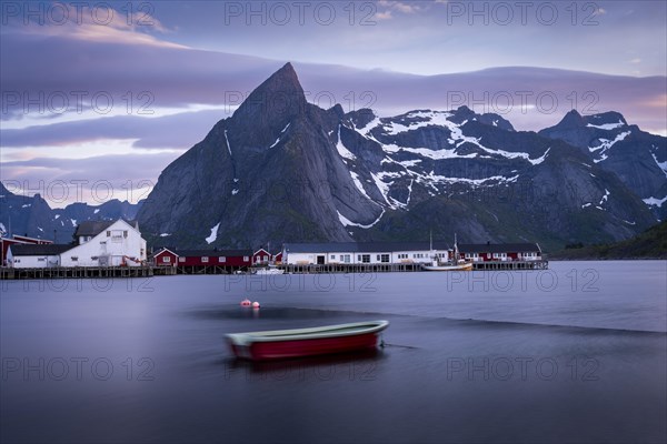 Landscape on the Lofoten Islands. The village of Hamnoy. A small boat in the foreground, blurred by long exposure. Houses and Mount Olstinden in the background. At night at the time of the midnight sun in good weather, some clouds in the sky. Early summer. Hamnoy, Moskenesoya, Lofoten, Norway, Europe