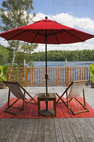 Red canvas cloth parasol and wooden folding canvas chairs on red mat on grey wood plank deck with railing overlooking lake with green forest in backyard in summer, Quebec, Canada, North America