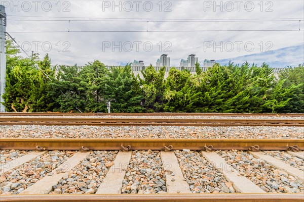Railway tracks on a bright day with lush green foliage and city buildings under a sky with fluffy clouds, in South Korea