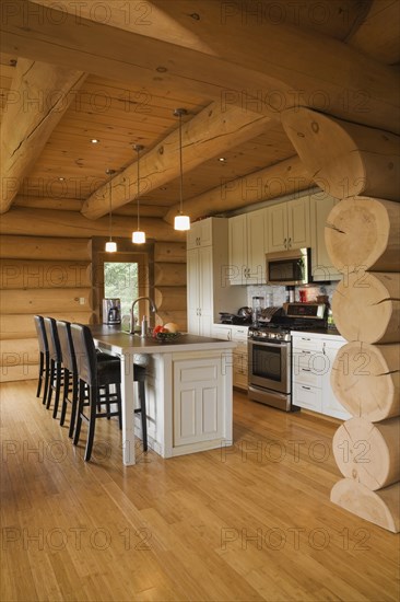 White painted wooden cabinets and island with dark brown wood and leather high back bar stools in kitchen with varnished floor boards inside luxurious contemporary Scandinavian style log cabin home, Quebec, Canada, North America