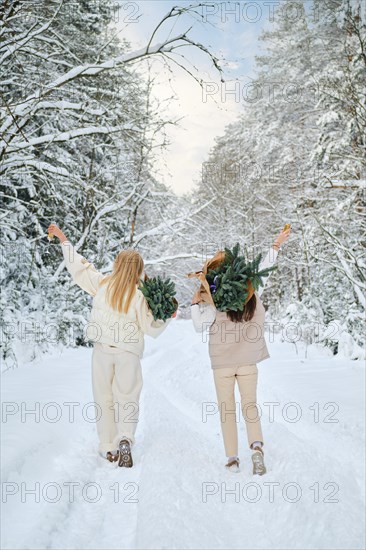 Rear view of two happy women walking in winter forest carrying small Christmas trees on their shoulders