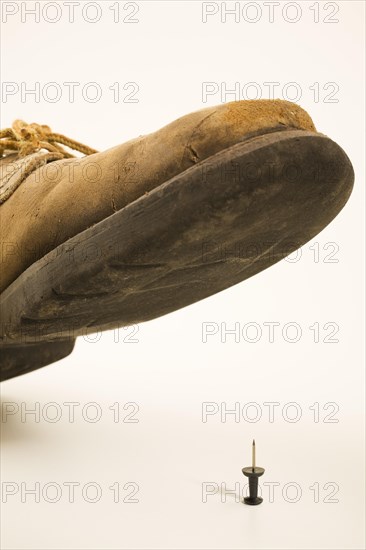 Close-up of elevated old and worn safety work shoe about to step on black upright pushpin with sharp point on white background, Studio Composition, Quebec, Canada, North America