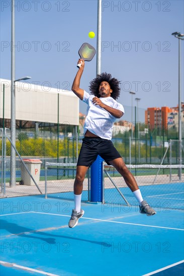 Vertical full length photo of a african american young sportive man jumping to reach the ball playing pickleball outdoors