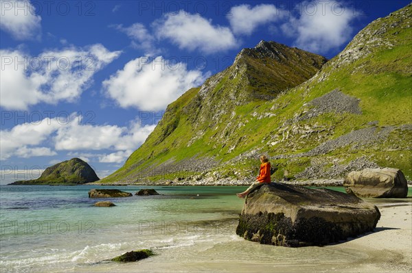 Landscape with sea at the sandy beach of Haukland (Hauklandstranda) with the mountain Veggen. A woman sits barefoot on a rock and looks out to sea. Shot during the day with blue sky, some clouds. Early summer. Haukland Beach, Haukland, Vestvagoya, Lofoten, Norway, Europe