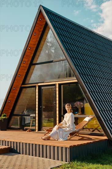 Mid adult woman in long sundress rests on wooden chair on terrace of tiny house in a sunny day