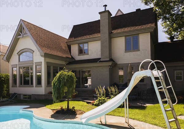 Rear view of a grey brick and cream cladding with grey trim contemporary cottage style home and landscaped backyard with in-ground swimming pool and slide in summer, Quebec, Canada, North America