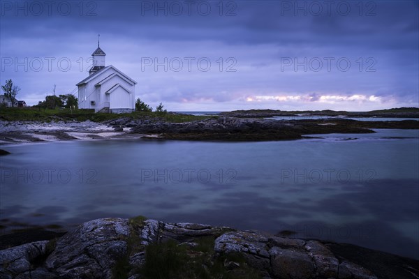 The white church of Gimsoy (Gimsoysand Church) by the sea, rocks in the foreground. At night at the time of the midnight sun. Cloudy sky, a narrow strip of light in the sky. Early summer. Long exposure. Gimsoya, Lofoten, Norway, Europe