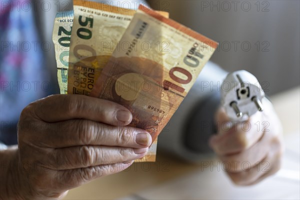 Senior citizen holding money and a power cable with plug in her hand at home, symbolising energy costs and poverty, Cologne, North Rhine-Westphalia, Germany, Europe