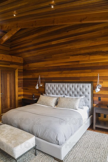 King size bed with grey upholstered panel headboard, ottoman and wooden end tables in master bedroom with grey nuanced rug and Ipe wood floor inside luxurious stained cedar and timber wood home, Quebec, Canada, North America