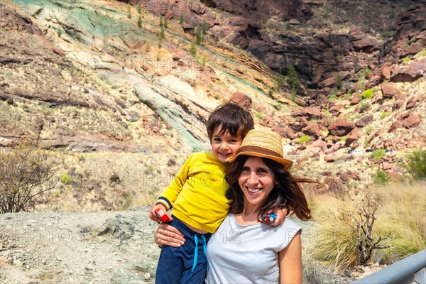 A woman and a child are posing for a picture in front of a rocky mountain. The woman is wearing a straw hat and the child is wearing a yellow shirt. Scene is lighthearted and joyful, as the mother