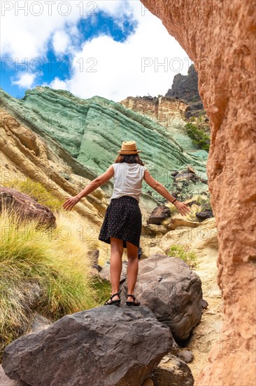 A tourist woman with a hat and open arms at the Azulejos de Veneguera or Rainbow Rocks in Mogan, Gran Canaria