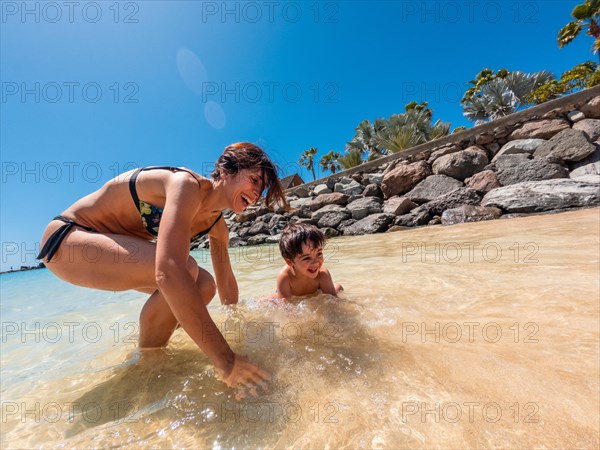 A woman and a child are playing in the ocean. The woman is helping the child to stand up in the water. Scene is joyful and playful