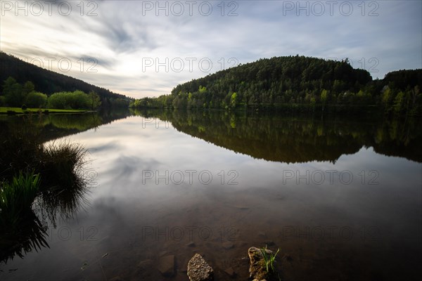 A lake in a landscape shot. A sunset and the natural surroundings are reflected in the water of the reservoir. Marbach reservoir, Odenwald, Hesse