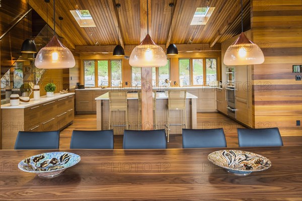 Illuminated industrial style copper with frosted glass pendant lighting fixtures over wooden dining table with black leather chairs plus kitchen with bamboo wood island and cabinets in background inside luxurious stained cedar and timber wood home with panoramic windows, Quebec, Canada, North America