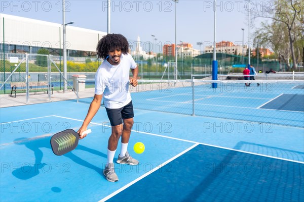 Full length photo of an smiling man with afro hair serving while playing pickleball in an outdoor court