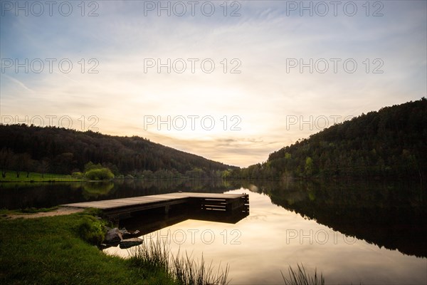 A lake in a landscape shot. A sunset and the natural surroundings are reflected in the water of the reservoir. Marbach reservoir, Odenwald, Hesse