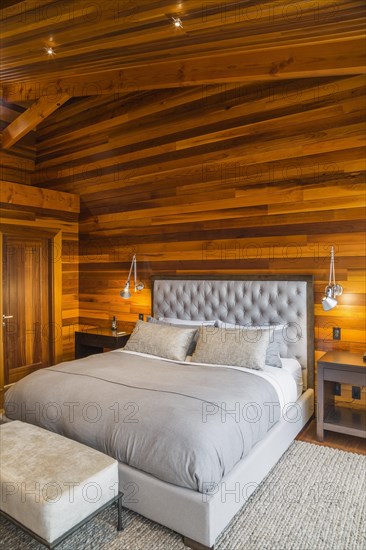 King size bed with grey upholstered panel headboard, ottoman and wooden end tables in master bedroom with grey nuanced rug and Ipe wood floor inside luxurious stained cedar and timber wood home, Quebec, Canada, North America