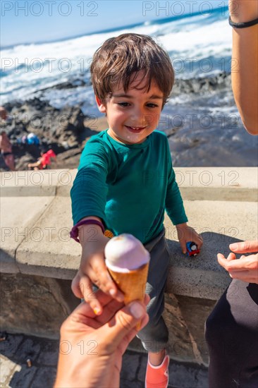 A young boy is holding an ice cream cone and giving it to another person. The scene is set on a beach, with the ocean in the background. Scene is lighthearted and playful