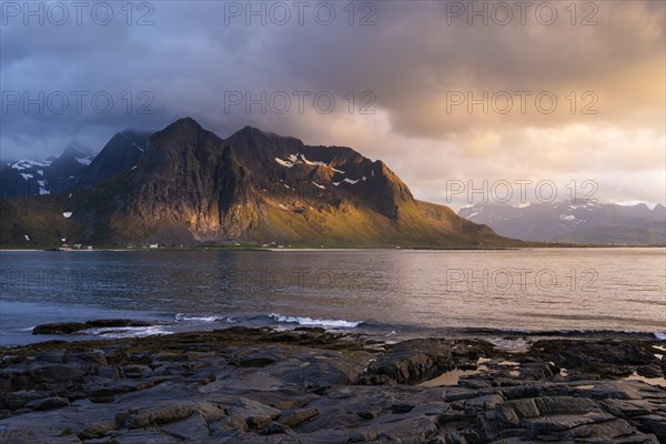 Landscape with sea and mountains on the Lofoten Islands, view across the fjord to the small town of Flakstad and the mountain Flakstadtinden as well as other mountains. Rocks in the foreground. At night at the time of the midnight sun. A few clouds in the sky, the sun shines sideways on the mountains. Golden hour. Early summer. Flakstadoya, Lofoten, Norway, Europe