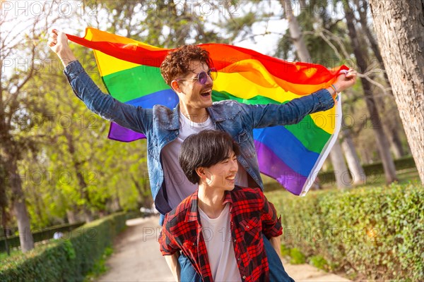 Multiracial gay couple waving lgbt flag piggybacking celebrating diversity in an urban park