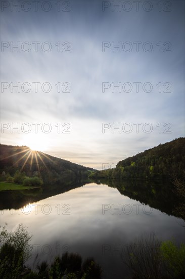 A lake in a landscape shot. A sunset and the natural surroundings are reflected in the water of the reservoir. Marbach reservoir, Odenwald, Hesse