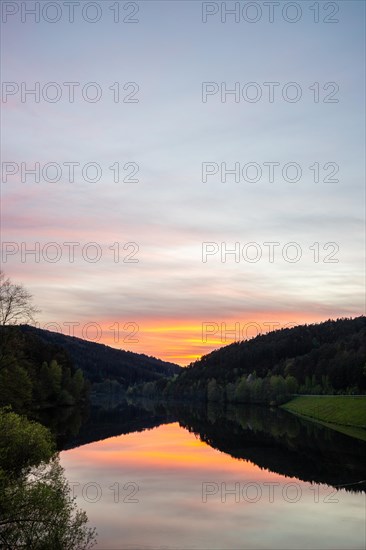 A lake in a landscape shot. A sunset and the natural surroundings are reflected in the water of the reservoir. Marbach reservoir, Odenwald, Hesse