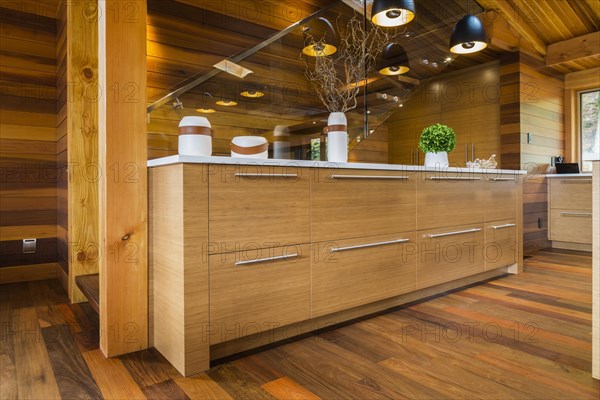 Bamboo wood buffet with white quartz countertop illuminated by black industrial style pendant lighting fixtures next to staircase with glass railing in kitchen with Ipe wood floor inside luxurious stained cedar and timber wood home, Quebec, Canada, North America